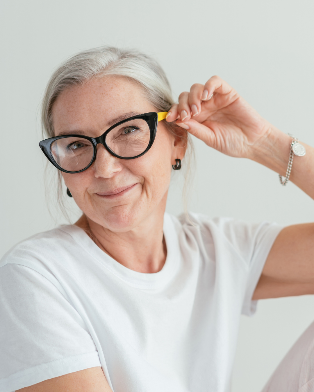 Smiling older woman with gray hair wears black glasses and a white shirt, adjusting the yellow temple of her glasses with her right hand.